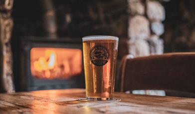 A photo of a pint of Gower Brewery Alcohol on a table in front of a wood burning fire in a pub.