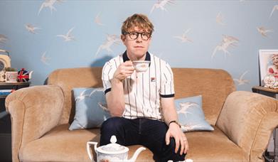 Photo of comedian Josh Widdicombe sitting on a sofa drinking tea