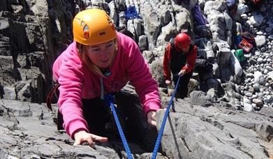 A lady wearing a pink jumper and orange hat taking part of the rock climbing activity available from Gower Adventures.