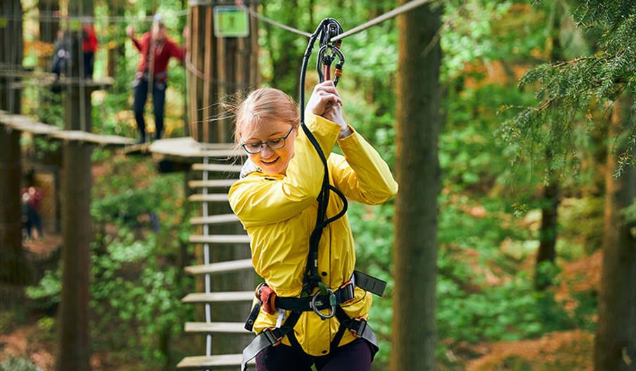 A woman in a yellow rain jacket climbing across a Go Ape Margam obstacle