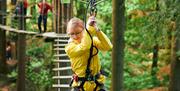 A woman in a yellow rain jacket climbing across a Go Ape Margam obstacle